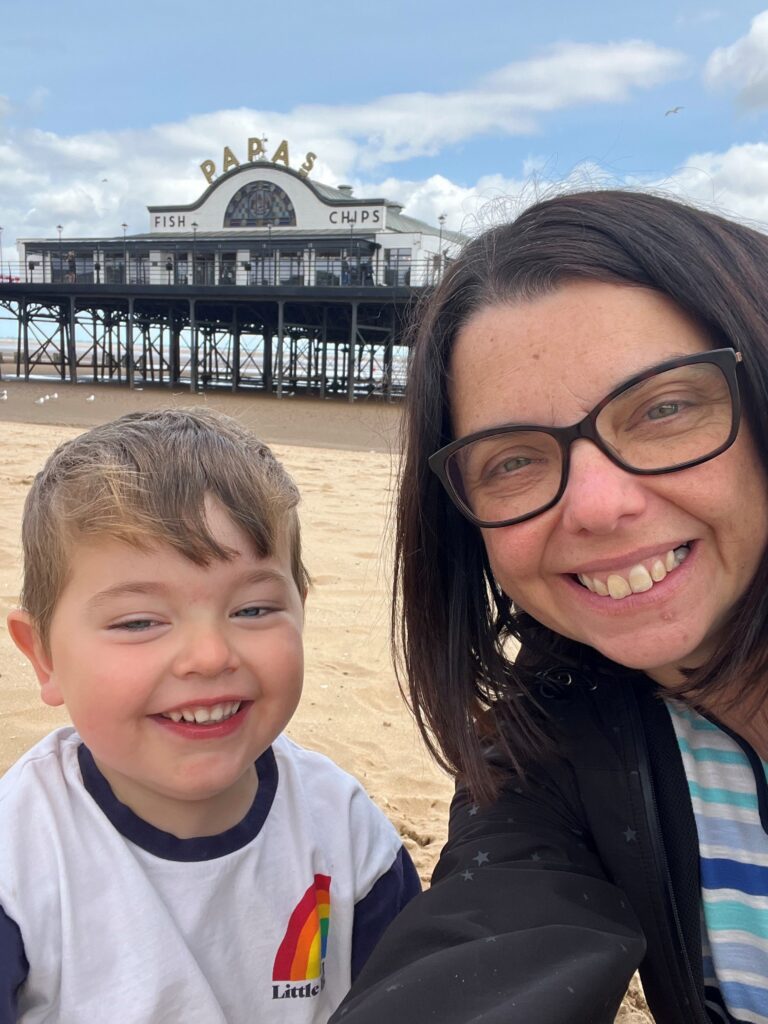 Cherylyn and her son, Oliver, are on the beach with a pier in the background. They smile at the camera and Papa's Fish and Chips can be seen.