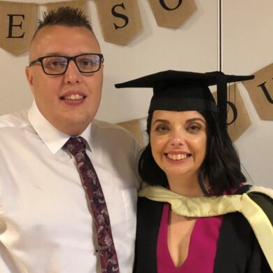 Cherylyn and her husband, Ryan, are at her graduation party. She wears a dark pink dress, with her graduation gown and cap on top. Ryan wears a white shirt, dark pink tie and black trousers. They both smile at the camera. A banner is behind them that says 