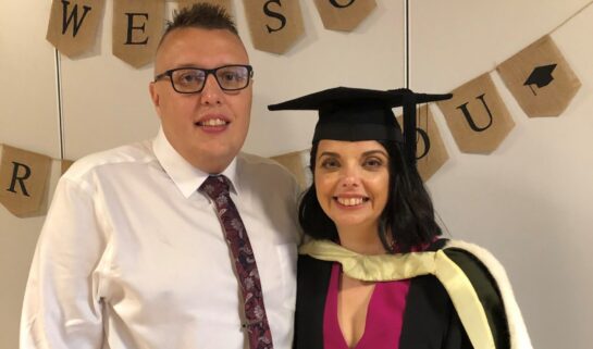 Cherylyn and her husband, Ryan, are at her graduation party. She wears a dark pink dress, with her graduation gown and cap on top. Ryan wears a white shirt, dark pink tie and black trousers. They both smile at the camera. A banner is behind them that says 
