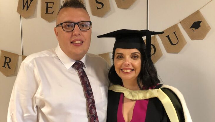 Cherylyn and her husband, Ryan, are at her graduation party. She wears a dark pink dress, with her graduation gown and cap on top. Ryan wears a white shirt, dark pink tie and black trousers. They both smile at the camera. A banner is behind them that says 