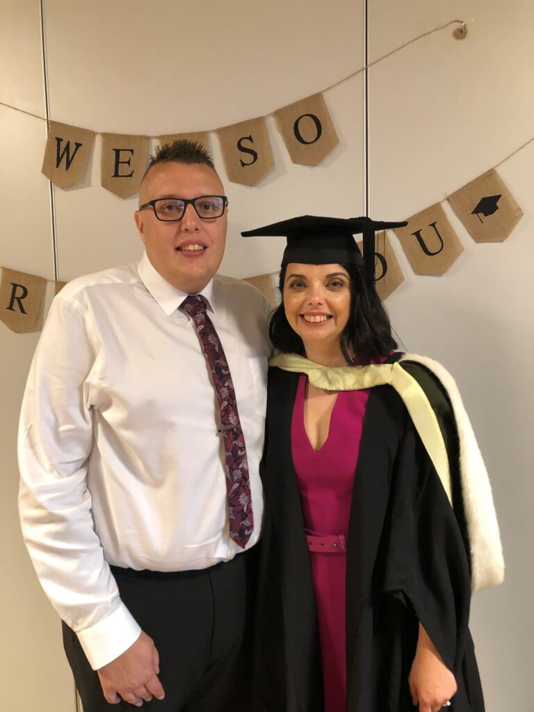 Cherylyn and her husband, Ryan, are at her graduation party. She wears a dark pink dress, with her graduation gown and cap on top. Ryan wears a white shirt, dark pink tie and black trousers. They both smile at the camera. A banner is behind them that says "we're so proud of you."