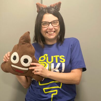 Cherylyn has short dark hair and black glasses. She is wearing a blue t-shirt that says Guts UK in green and white. She is wearing poo boppers on her head and is holding a poo cushion with eyes and a smiley face.