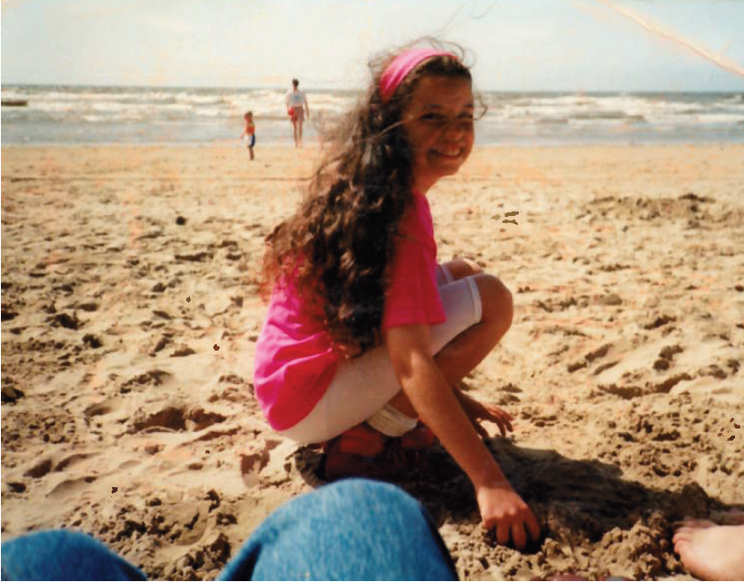 Cherylyn is on the beach when she was younger. She wears white cycle shorts, a pink t-shirt and has long, wavy brown hair. She smiles at the camera, and also has a pink headband that matches her t-shirt.