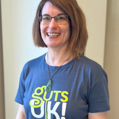 Chrissy is wearing a navy Guts UK t-shirt. She stands with a cream cupboard in the background, and wears glasses and a necklace. She has her hair styled in a bob.