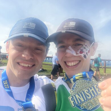 A selfie of Paul and Danielle smiling after completing the Great North Run. Both are wearing running caps and proudly holding up their "Finisher 13.1 Miles" medals. Danielle is wearing a green Macmillan Cancer Support shirt and has a feeding tube secured to her cheek with medical tape.
