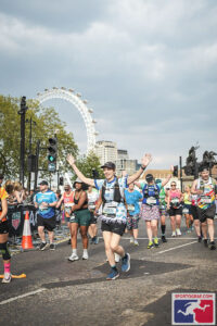 Danielle runs with her arms up at the London Marathon, with the London eye in the background. She is wearing a blue running jersey, black shorts, black cap and a hydration vest. She is surrounded by other runners.