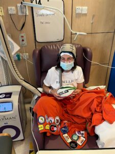 Danielle sits on a purple hospital chair and is wearing a white t-shirt, a blue surgical mask and a Paxman scalp cooling cap. An orange blanket adorned with merit patches covers the bottom half of her body and she is holding a Six Nations rugby ball. A Paxman scalp cooling machine stands to her left.
