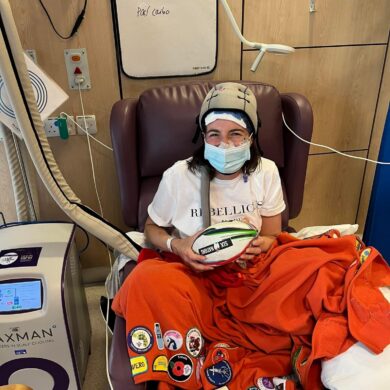 Danielle sits on a purple hospital chair and is wearing a white t-shirt, a blue surgical mask and a Paxman scalp cooling cap. An orange blanket adorned with merit patches covers the bottom half of her body and she is holding a Six Nations rugby ball. A Paxman scalp cooling machine stands to her left.