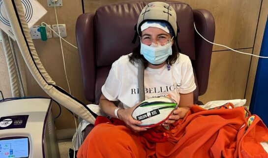 Danielle sits on a purple hospital chair and is wearing a white t-shirt, a blue surgical mask and a Paxman scalp cooling cap. An orange blanket adorned with merit patches covers the bottom half of her body and she is holding a Six Nations rugby ball. A Paxman scalp cooling machine stands to her left.