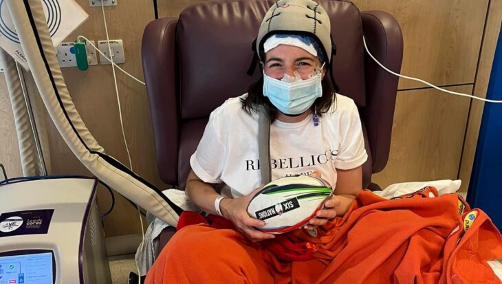 Danielle sits on a purple hospital chair and is wearing a white t-shirt, a blue surgical mask and a Paxman scalp cooling cap. An orange blanket adorned with merit patches covers the bottom half of her body and she is holding a Six Nations rugby ball. A Paxman scalp cooling machine stands to her left.