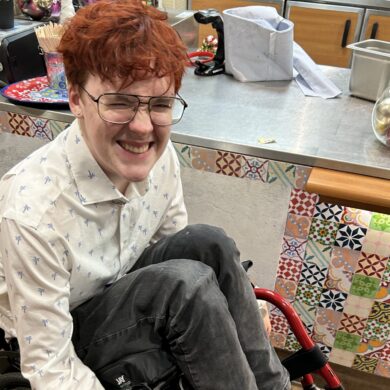 A smiling Griffin sitting in a red manual wheelchair, wearing a white button-down shirt and black denim jeans.