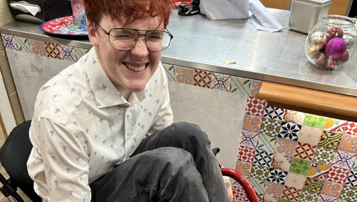 A smiling Griffin sitting in a red manual wheelchair, wearing a white button-down shirt and black denim jeans.