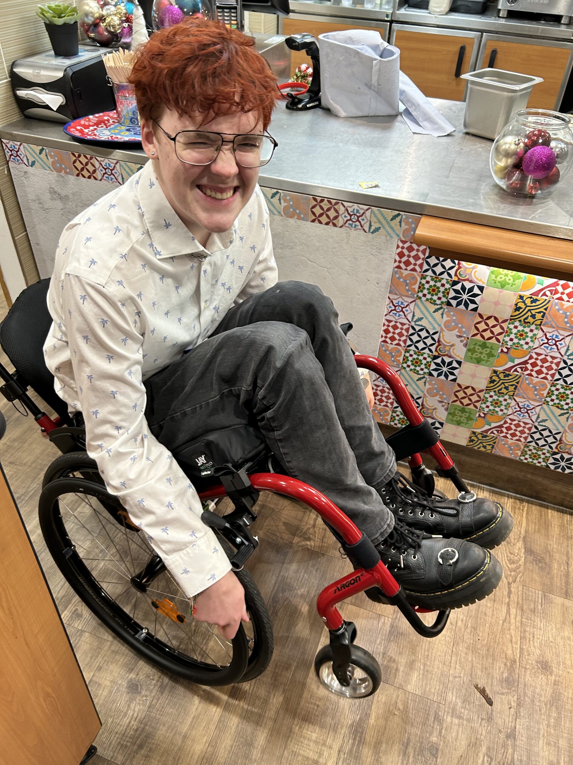 A smiling Griffin sitting in a red manual wheelchair, wearing a white button-down shirt and black boots.