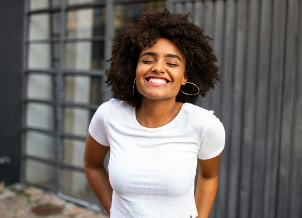 Gemini said A young woman with curly hair smiles brightly with her eyes closed, standing outdoors in front of a building with large windows.