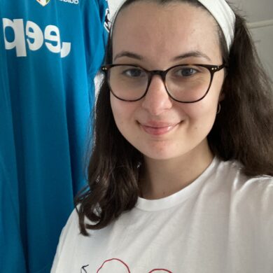 A close-up selfie of Sophia wearing a white headband, glasses, and a white t-shirt, with a blue football top hanging behind her.