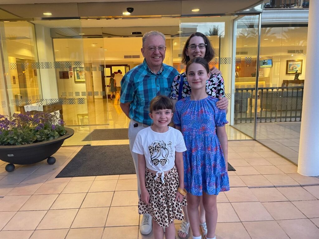 A group photo taken at dusk in front of a hotel entrance with glass doors. Leslie, wearing a teal and navy checkered shirt, stands in the back left. Beside him is a woman in a black floral top. In front of them are two girls: one wearing a white t-shirt and a leopard-print skirt, and the other in a blue floral dress. They are all smiling at the camera on a tiled patio.