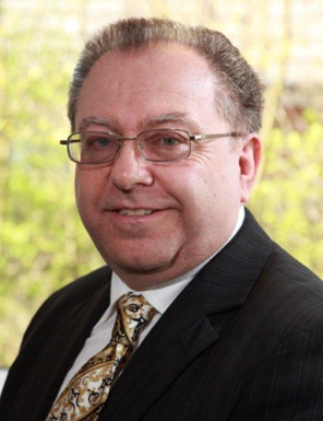 A portrait of Leslie, a man with short brown hair and glasses, wearing a dark pinstripe suit, a white shirt, and a gold and white patterned tie. He is smiling slightly at the camera against a blurred background of green foliage.