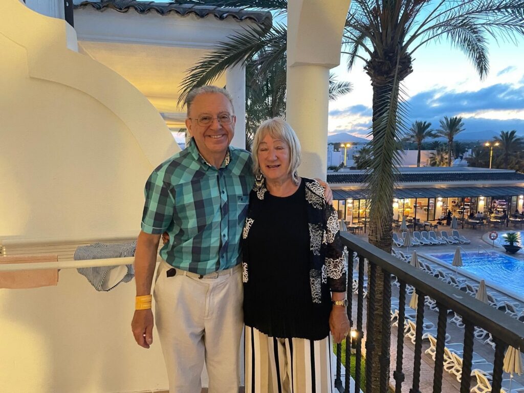 Leslie and his wife stand close together on a balcony during the evening, smiling warmly at the camera. Leslie, on the left wears a teal and navy checkered short-sleeved shirt and light-coloured trousers, while the woman on the right wears a black top with a white lace-patterned cardigan and striped trousers. Behind them, a resort setting is visible, featuring palm trees, a swimming pool with lounge chairs, and buildings under a soft, twilight sky.