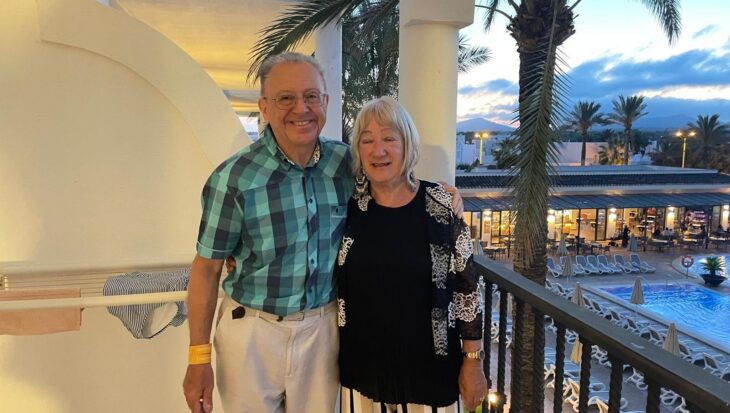 Leslie and his wife stand close together on a balcony during the evening, smiling warmly at the camera. Leslie, on the left wears a teal and navy checkered short-sleeved shirt and light-coloured trousers, while the woman on the right wears a black top with a white lace-patterned cardigan and striped trousers. Behind them, a resort setting is visible, featuring palm trees, a swimming pool with lounge chairs, and buildings under a soft, twilight sky.