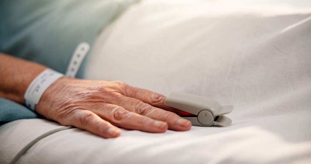 A hand resting on a hospital bed wearing a white identification wristband and a fingertip pulse oximeter for oxygen saturation monitoring.