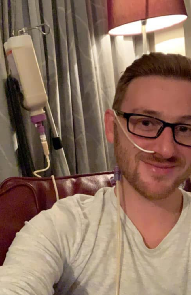 A close-up selfie of Toby, a man with short brown hair, a beard, and black-rimmed glasses, smiling at the camera. He has a white nasogastric (NG) tube inserted into his right nostril, which is secured to his cheek and connected to a larger tube running down his chest. In the background, a bottle containing a beige liquid (enteral feed) hangs from a metal stand, connected to the tubing. Toby is wearing a light-coloured long-sleeved shirt and is sitting in a dark red chair.
