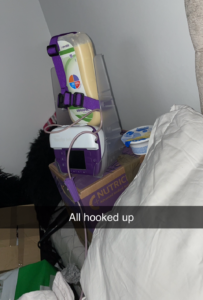 A close-up shot of an enteral feeding station labelled with the text "All hooked up." A large bag of beige liquid formula is held upright in a clear plastic stand by a purple strap. Below the bag sits a purple and white electronic feeding pump with a digital screen. The entire setup is resting on a cardboard box next to a bed with white linens.