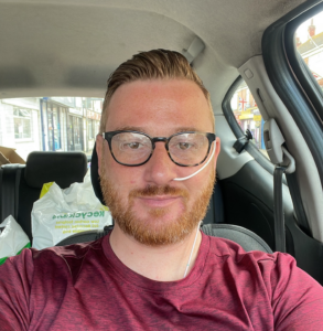 Toby, with short reddish-brown hair, glasses, and a beard, is sitting in the passenger seat of a car. He is wearing a maroon t-shirt and has a thin, white nasogastric (NG) tube inserted into his left nostril, secured to his cheek with clear adhesive. He is looking directly at the camera with a slight smile. In the background, there are white plastic shopping bags on the back seat and a street view through the car window.