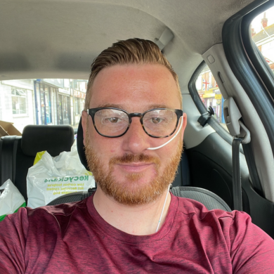 Toby, with short reddish-brown hair, glasses, and a beard, is sitting in the passenger seat of a car. He is wearing a maroon t-shirt and has a thin, white nasogastric (NG) tube inserted into his left nostril, secured to his cheek with clear adhesive. He is looking directly at the camera with a slight smile. In the background, there are white plastic shopping bags on the back seat and a street view through the car window.