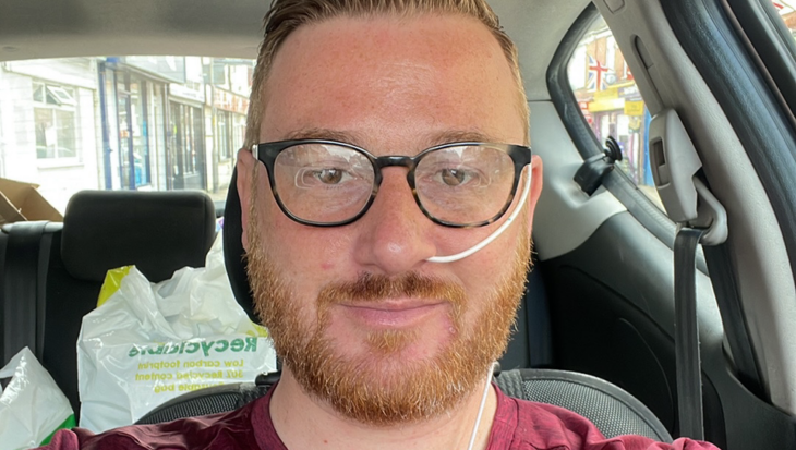 Toby, with short reddish-brown hair, glasses, and a beard, is sitting in the passenger seat of a car. He is wearing a maroon t-shirt and has a thin, white nasogastric (NG) tube inserted into his left nostril, secured to his cheek with clear adhesive. He is looking directly at the camera with a slight smile. In the background, there are white plastic shopping bags on the back seat and a street view through the car window.