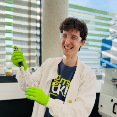 A photograph of a researcher in a lab, wearing a white lab coat and green gloves.