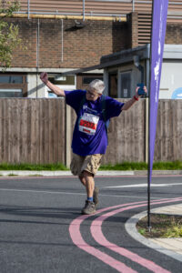 Ceri is mid-stride during an outdoor event, wearing a purple t-shirt with a race bib. She has her arms raised in celebration. She is wearing cargo shorts and hiking boots.