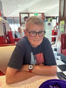 Ceri is sitting at a booth in a diner. She is wearing a dark t-shirt and a watch with a red strap, resting her arms on a table with a red-and-white patterned top.