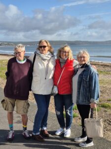 Ceri is standing with her friends on a paved path next to a beach. She is wearing a purple jumper, tan cargo shorts, and patterned socks with trainers. The group is smiling with a calm sea and hills in the background under a cloudy sky.