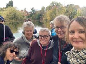 Ceri is in the centre with her four other friends outdoors. She is wearing a purple hoodie. Most of the group members are wearing headsets with microphones. A lake and autumn trees are visible behind them.