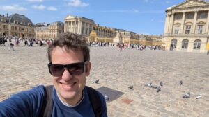 David smiling in a selfie at a large, historic cobblestone courtyard, with the palace of Versailles and pigeons in the background.