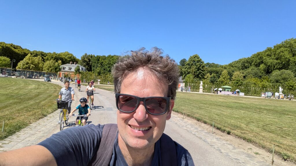 A selfie of David smiling on a sunny day while cycling on a paved path, with other cyclists and lush green trees in the background.