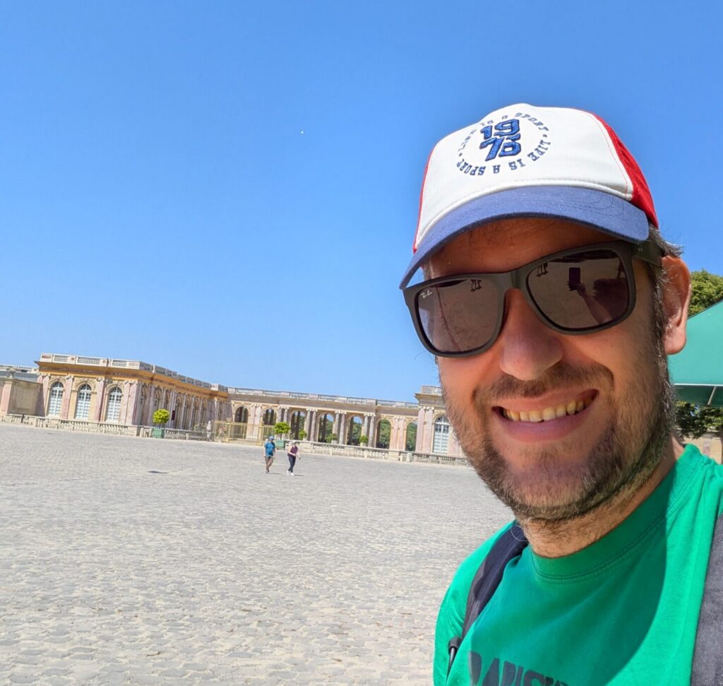 A close-up selfie of David wearing a green t-shirt and a baseball cap, standing in front of a wide cobblestone plaza and the Palace of Versailles.