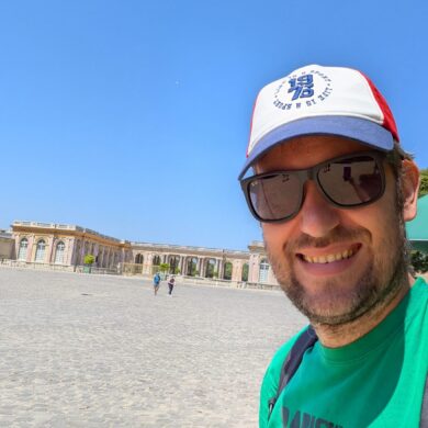 A close-up selfie of David wearing a green t-shirt and a baseball cap, standing in front of a wide cobblestone plaza and the Palace of Versailles.