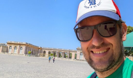 A close-up selfie of David wearing a green t-shirt and a baseball cap, standing in front of a wide cobblestone plaza and the Palace of Versailles.