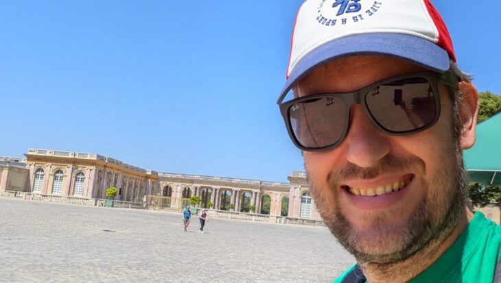 A close-up selfie of David wearing a green t-shirt and a baseball cap, standing in front of a wide cobblestone plaza and the Palace of Versailles.