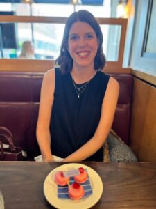 Grace has shoulder-length brown hair and smiles at a restaurant table, with a plate of three pink macarons in the foreground.