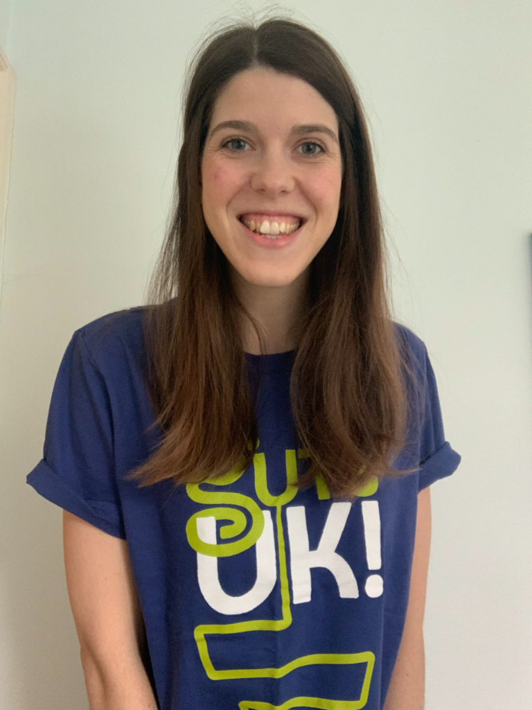 Grace has long, straight brown hair and is smiling, wearing a blue t-shirt that features the text "GUTS UK!" in white and lime green.