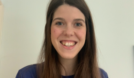 Grace has long, straight brown hair and is smiling, wearing a blue t-shirt that features the text 