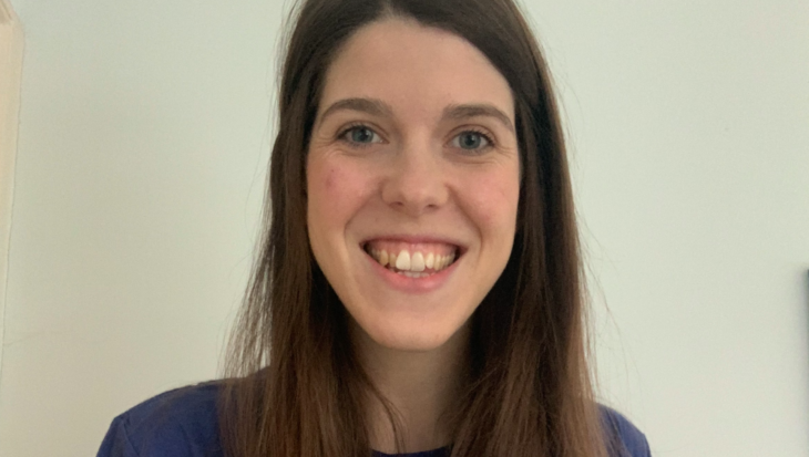Grace has long, straight brown hair and is smiling, wearing a blue t-shirt that features the text 
