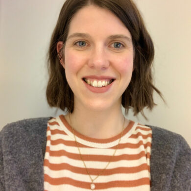 A close-up portrait of Grace, smiling, with a wavy brown bob, wearing a t-shirt with peach and white horizontal stripes, a grey cardigan, and a thin, gold necklace.