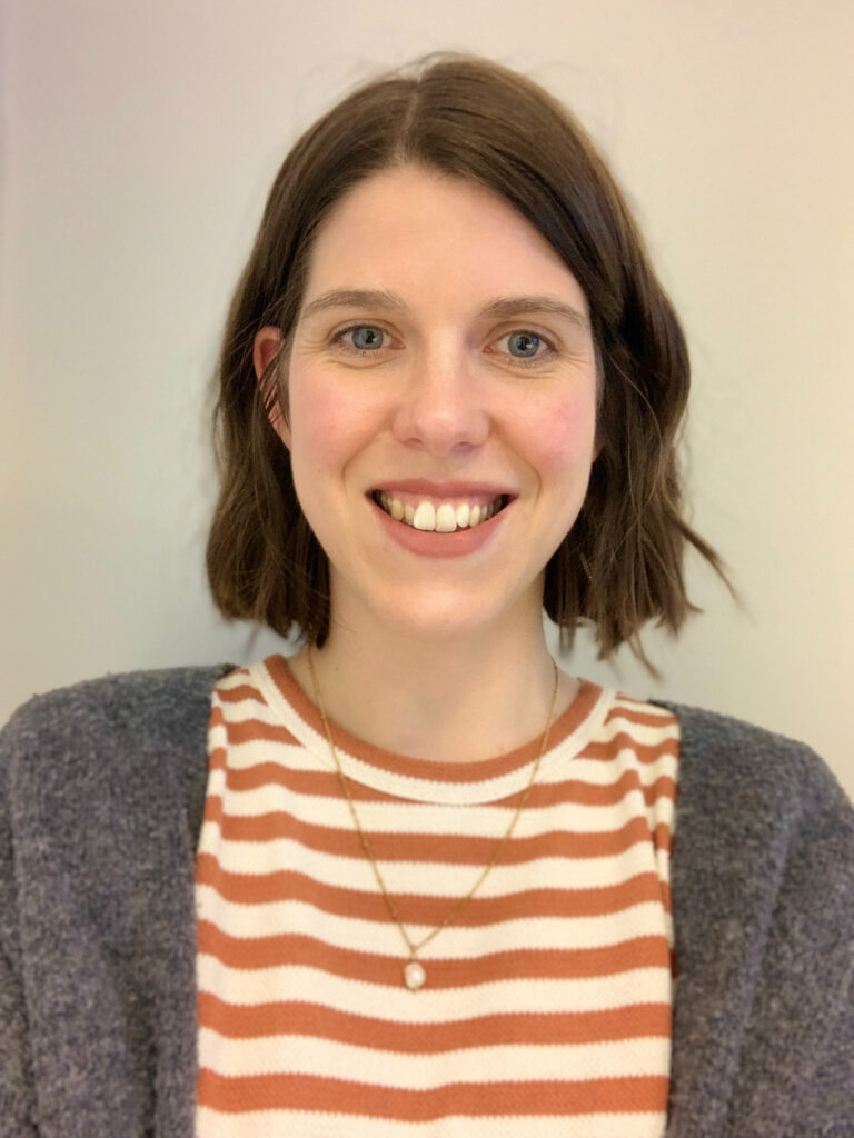 A close-up portrait of Grace, smiling, with a wavy brown bob, wearing a t-shirt with peach and white horizontal stripes, a grey cardigan, and a thin, gold necklace.