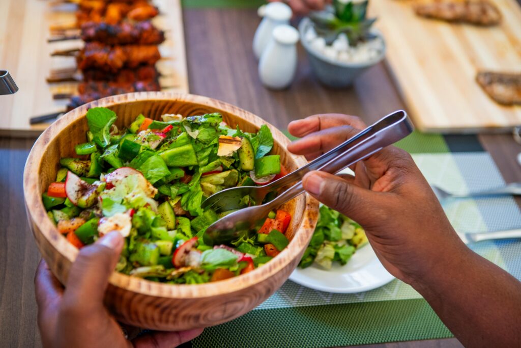 
A person's hands using metal tongs to serve a fresh green salad with radishes and cucumbers from a wooden bowl. In the background, skewers of grilled meat and small condiment containers are visible on a wooden table.