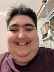 A cheerful, high-angle close-up selfie of Morgan smiling broadly. They are wearing a maroon t-shirt. The photo captures a joyful expression, with their short dark hair styled upward. In the background.