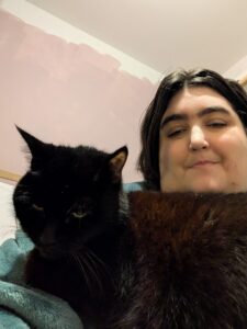 A close-up selfie of Morgan and a black cat. Morgan is smiling gently at the camera, while a large, fluffy black cat rests on their chest. The background is simple, showing a wall with light pink and white paint. The lighting is soft, creating a cozy and personal atmosphere.