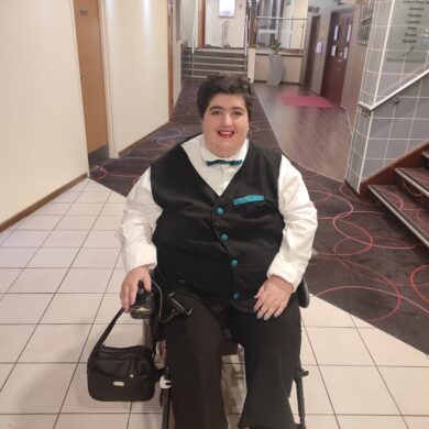 A full-length photo of Morgan smiling in a brightly lit hallway. They are sitting in a power wheelchair, dressed formally in a white collared shirt, a black waistcoat with teal buttons and a teal pocket square, a teal bowtie, and black trousers. A small black bag is attached to the side of the wheelchair. The background shows a hotel-style corridor with neutral-coloured walls, a patterned carpet, and a staircase.